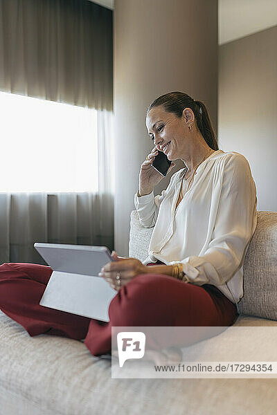 Lächelnde weibliche Fachkraft  die mit einem Smartphone spricht und mit einem digitalen Tablet auf dem Sofa sitzt