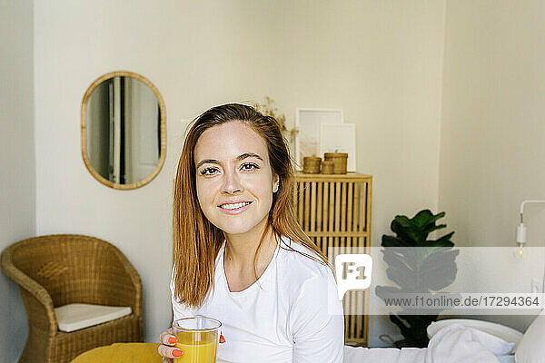 Lächelnde Frau trinkt Saft  während sie zu Hause auf dem Bett sitzt