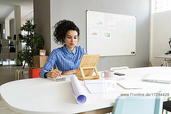 Junge Geschäftsfrau schreibt in einen Notizblock  während sie ein digitales Tablet im Büro benutzt