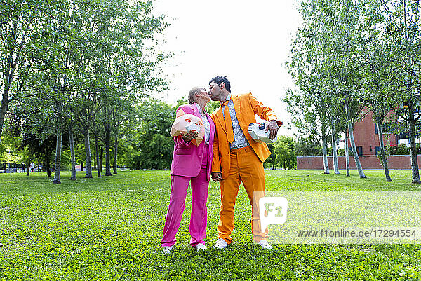Erwachsenes Paar in lebhaften Anzügen küsst sich im öffentlichen Park mit Tiermasken in den Händen