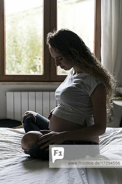 Junge schwangere Frau betrachtet ihren Bauch  während sie auf dem Bett sitzt