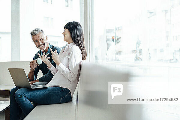 Lächelnde Geschäftsfrau sitzt mit Laptop neben einem Kollegen in einem Café
