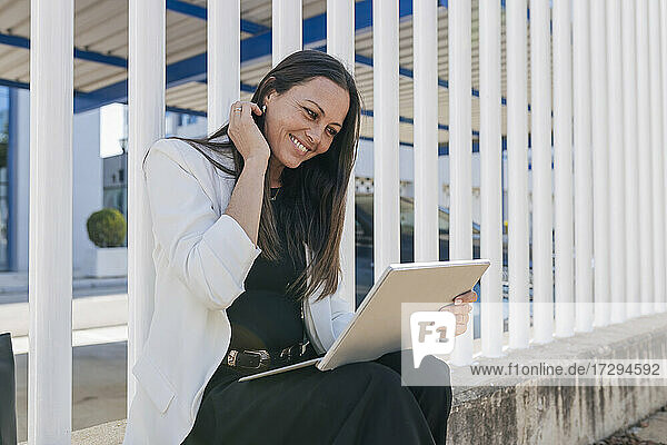 Lächelnde Geschäftsfrau  die ein digitales Tablet benutzt  während sie vor einem Zaun sitzt