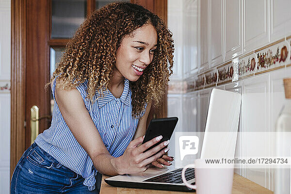 Lächelnde Frau mit lockigen Haaren und Mobiltelefon  die zu Hause einen Laptop benutzt