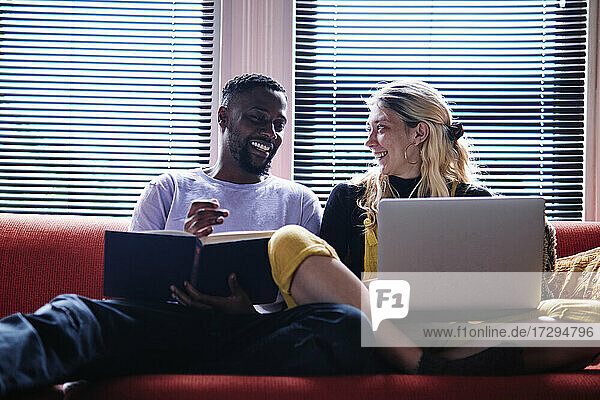 Lächelndes Paar mit Buch und Laptop im Gespräch auf dem Sofa sitzend