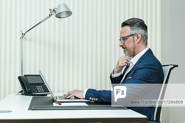Männlicher Unternehmer mit Hand am Kinn  der einen Laptop am Schreibtisch benutzt