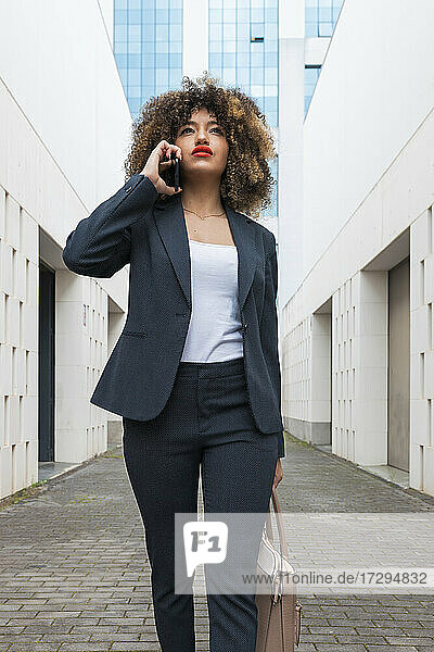 Geschäftsfrau mit Handtasche  die beim Spazierengehen inmitten von Bürogebäuden mit ihrem Smartphone telefoniert