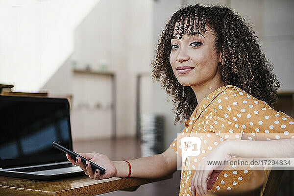 Junge Freiberuflerin hält ihr Smartphone in der Hand  während sie in einem Café am Laptop sitzt