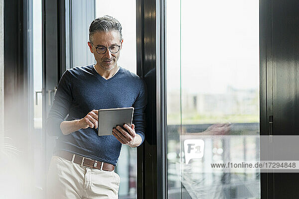 Male entrepreneur using digital tablet while leaning on glass window in office