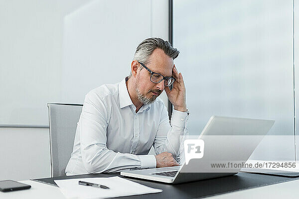 Geschäftsmann mit Kopf in der Hand sitzt mit Laptop am Schreibtisch