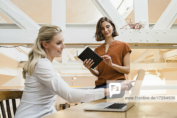 Young female professional looking at coworker pointing on laptop while discussing plan in creative office