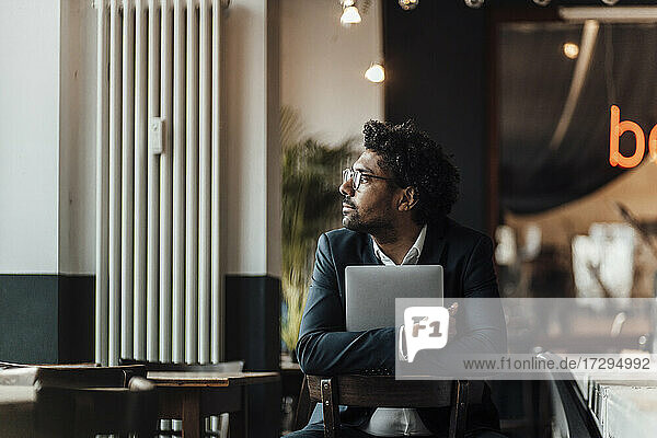 Male entrepreneur holding laptop while sitting on chair in cafe looking away