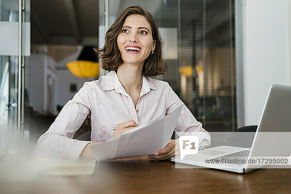 Beautiful smiling female entrepreneur sitting with document at desk while looking away