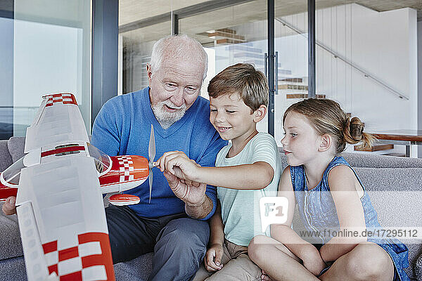 Senior man holding toy airplane while sitting with grandchildren in living room at home