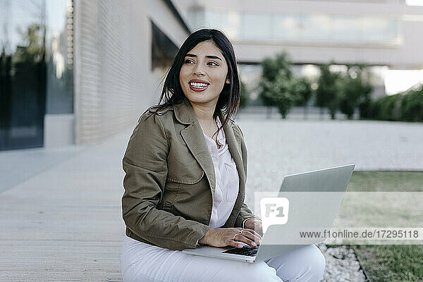 Young businesswoman with laptop smiling while looking away on footpath