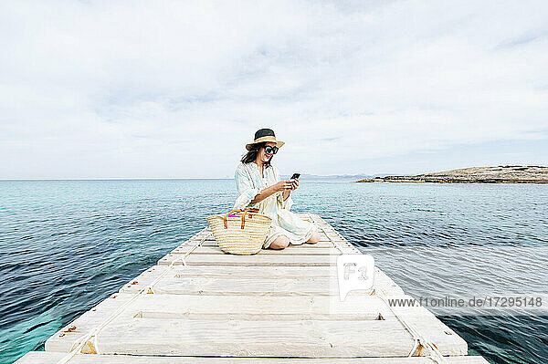 Lächelnde Frau  die ein Mobiltelefon benutzt  während sie im Urlaub auf einem Steg sitzt