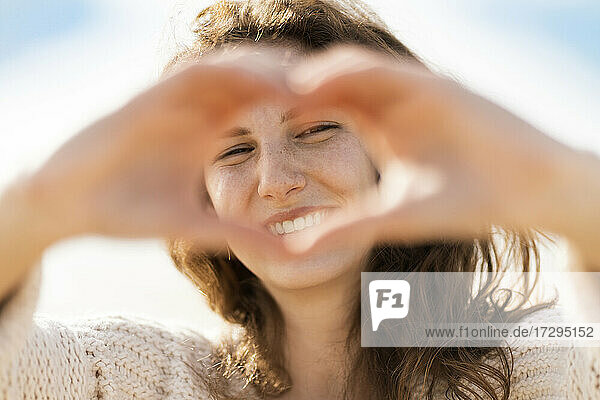 Glückliche junge Frau  die durch ein von Hand geformtes Herz am Strand während eines sonnigen Tages schaut
