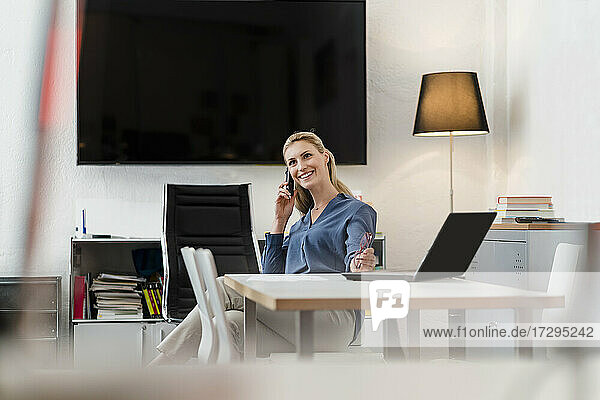 Glückliche weibliche Fachkraft  die am Schreibtisch im Büro sitzt und mit ihrem Smartphone spricht