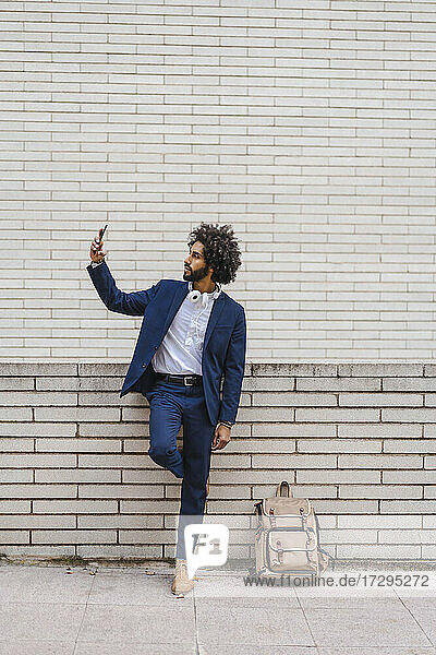Geschäftsmann  der ein Selfie mit seinem Mobiltelefon macht  während er mit seinem Rucksack auf dem Fußweg steht