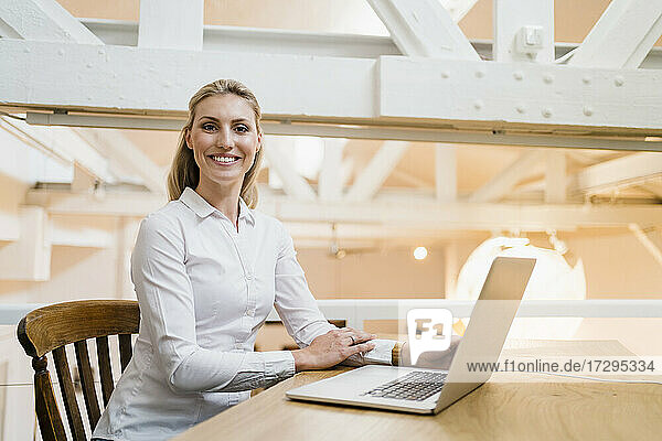 Smiling blond female entrepreneur sitting with laptop in creative office