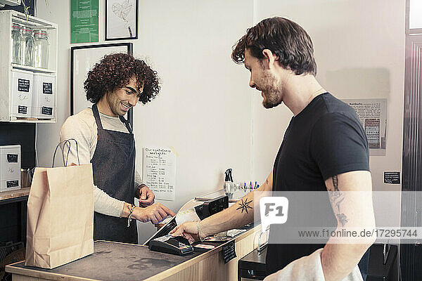 Lächelnde Kassiererin mit Registrierkasse  während sie mit einem männlichen Kunden an der Kasse eines Bioladens steht