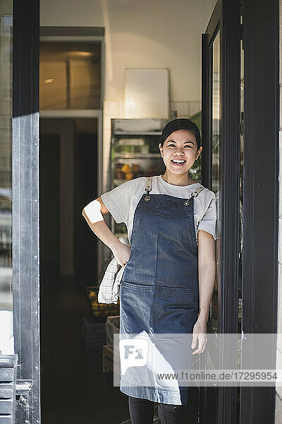 Porträt einer lächelnden Besitzerin mit Hand auf der Hüfte  die am Eingang eines Geschäfts steht