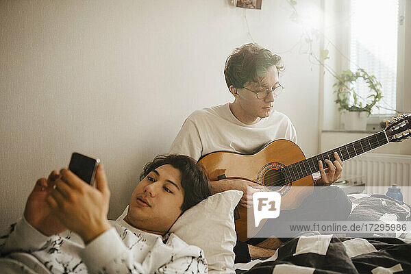 Young man using smart phone while male friend playing guitar at home