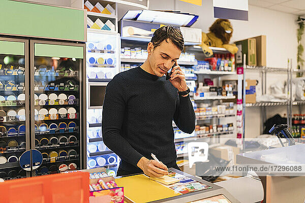 Männlicher Ladenbesitzer schreibt  während er an der Kasse mit dem Handy spricht