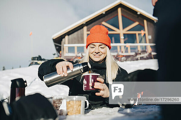 Lächelnde Frau gießt Kaffee in Tasse während des Winters