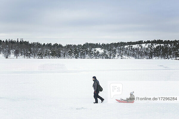 Mann in voller Länge  der seine Tochter beim Winterspaziergang auf dem Schlitten sitzend zieht