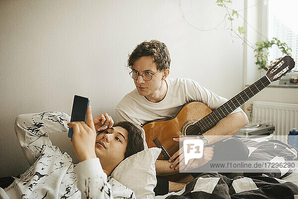 Man with guitar peeking while male friend using smart phone in bedroom