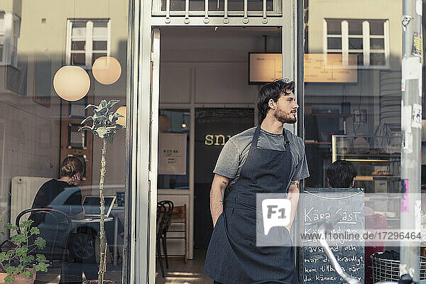 Männlicher Besitzer  der wegschaut  während er mit den Händen in den Taschen am Eingang eines Cafés steht