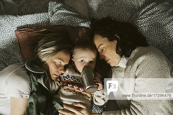 Mädchen trinkt Milch und liegt inmitten von lesbischen Müttern auf dem Bett zu Hause