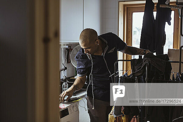 Glatzköpfiger Mann schaut auf die Waschmaschine  während er im Hauswirtschaftsraum Wäsche wäscht
