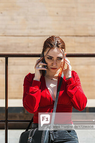 Junge rothaarige Frau  die in einem städtischen Raum mit einem Mobiltelefon spricht. Bekleidet mit einer roten Bluse.