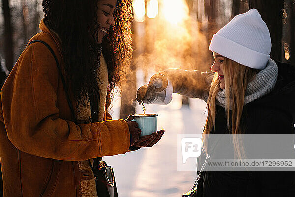 Glückliche Frau gießt Tee für ihre Freundin ein  die im Winter im Park steht
