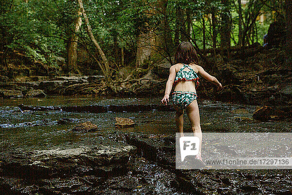 Rückansicht eines kleinen Kindes  das in einem sonnenbeschienenen Wald durch einen Bach läuft