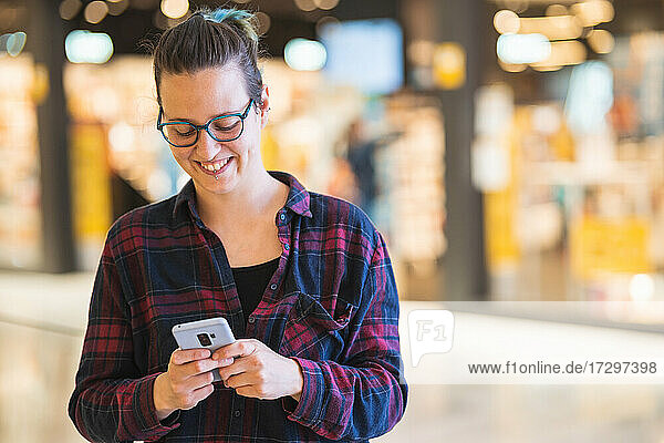 Spanish woman smiling and texting on smartphone while shopping in mall