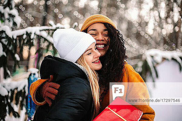 Glückliche Freundinnen umarmen sich und halten ein Weihnachtsgeschenk am Weihnachtsbaum