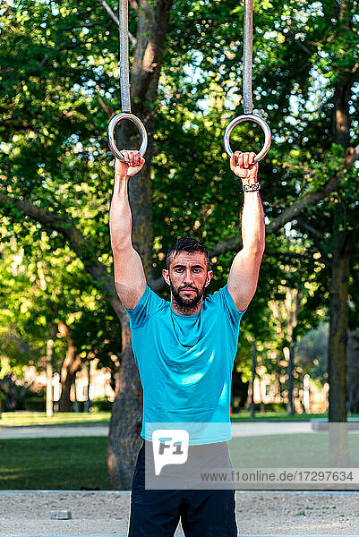 Dark-haired athlete with beard holding on to gymnastic rings.