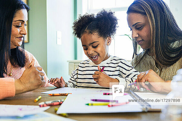 Mutter und Großmutter mit fröhlichem Mädchen  das mit Buntstiften auf Papier zu Hause zeichnet