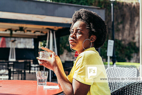 Porträt eines schwarzen Mädchens mit Afrofrisur und Ohrringen  das auf der Terrasse einer Bar Kaffee und ein Glas Wasser trinkt.