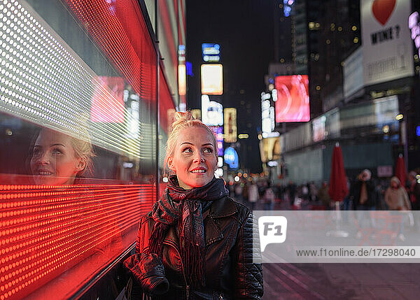 Cheerful woman standing near illuminated building wall in evening city