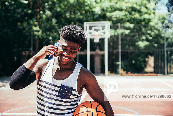 Porträt eines schwarzen afroamerikanischen Jungen  der auf einem städtischen Basketballplatz mit seinem Mobiltelefon spricht.