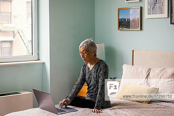 Ältere Frau benutzt einen Laptop  während sie im Schlafzimmer zu Hause sitzt