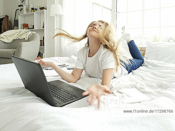 junge Frau spielt mit langen blonden Haaren auf dem Bett liegend mit Laptop