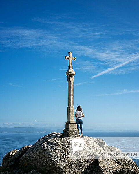 Frau auf einem Aussichtsfelsen mit einer Kreuzskulptur in Finisterre