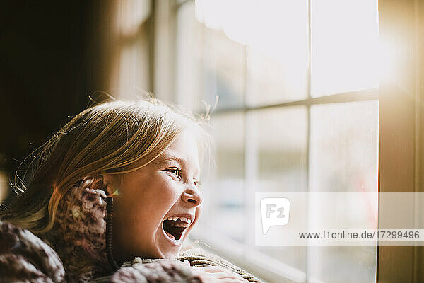 Junges Mädchen mit blonden Haaren lachend vor einem Fenster mit Sonneneinstrahlung