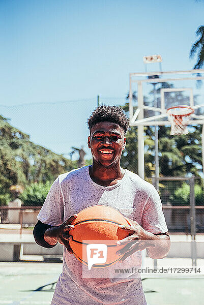 Porträt eines schwarzen afroamerikanischen Jungen  der lächelt und auf einem städtischen Basketballplatz einen Basketball gegen seine Brust hält.