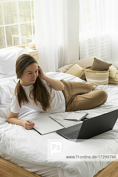 Woman in white T-shirt lies on bed and looking at laptop